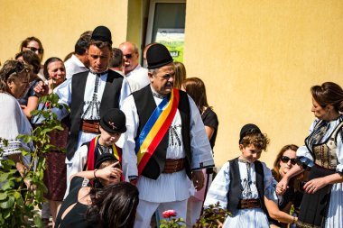 Geleneksel siyah motiflerle dikilmiş ulusal çoban elbisesi giymiş, Romanya 'da bir belediye başkanı, yuvarlak siyah bir şapka, geniş bir deri kimir ve belediye başkanı için üç renkli bir eşarp giyiyor.