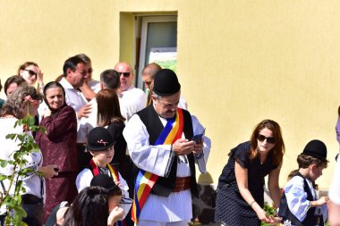 Geleneksel siyah motiflerle dikilmiş ulusal çoban elbisesi giymiş, Romanya 'da bir belediye başkanı, yuvarlak siyah bir şapka, geniş bir deri kimir ve belediye başkanı için üç renkli bir eşarp giyiyor.