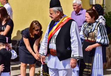 Geleneksel siyah motiflerle dikilmiş ulusal çoban elbisesi giymiş, Romanya 'da bir belediye başkanı, yuvarlak siyah bir şapka, geniş bir deri kimir ve belediye başkanı için üç renkli bir eşarp giyiyor.