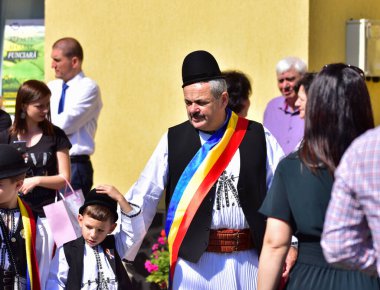 Geleneksel siyah motiflerle dikilmiş ulusal çoban elbisesi giymiş, Romanya 'da bir belediye başkanı, yuvarlak siyah bir şapka, geniş bir deri kimir ve belediye başkanı için üç renkli bir eşarp giyiyor.