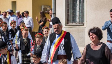 Geleneksel siyah motiflerle dikilmiş ulusal çoban elbisesi giymiş, Romanya 'da bir belediye başkanı, yuvarlak siyah bir şapka, geniş bir deri kimir ve belediye başkanı için üç renkli bir eşarp giyiyor.
