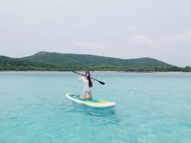  Sörf tahtasında dinlenen, turkuaz deniz suyunda yüzen bir kadın. Ayağa kalk Paddle bordası. Yaz eğlencesi, tatil seyahati. Etkin yaşam biçimi