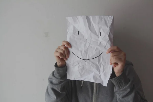 Woman holding a smiling face emoticon in front of face against a gray background, depression, mental health, masking feelings.
