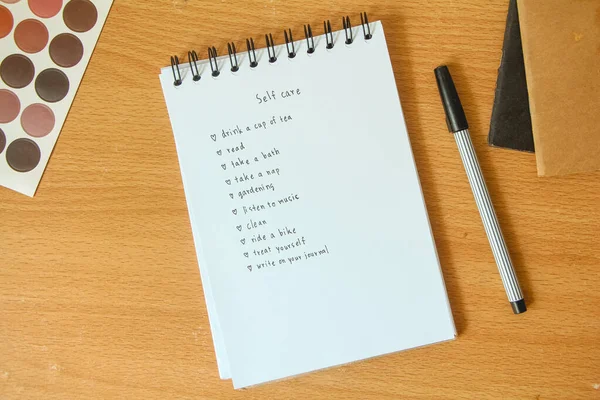 Text list for self care on notebook with pen on white background, office desk.