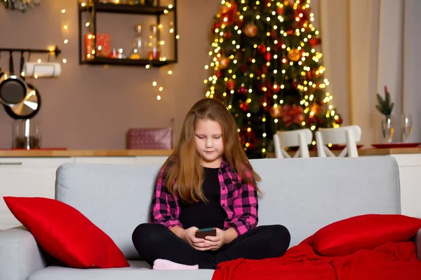 彼女の手のビデオ通話チャットでスマートフォンを持つソファの上のかわいい女の子は陽気なクリスマスを祝います クリスマス ツリー ボケのために飾られたリビングルームとキッチン オンラインクリスマスの挨拶の概念 — ストック写真