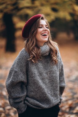 Sonbahar parkında eğlenen neşeli genç bir kadının portresi. Sonbahar kırmızı bereli ve gri kazaklı güzel sarışın kız. Doğada rahatla.