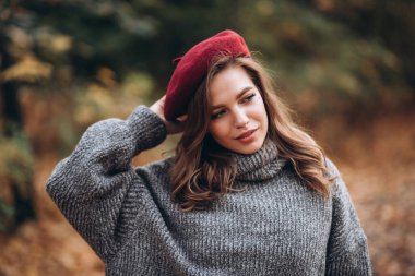 Sonbahar parkında eğlenen neşeli genç bir kadının portresi. Sonbahar kırmızı bereli ve gri kazaklı güzel sarışın kız. Doğada rahatla.