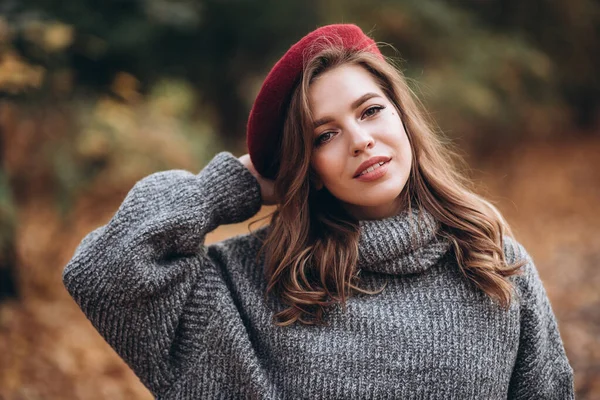 Sonbahar parkında eğlenen neşeli genç bir kadının portresi. Sonbahar kırmızı bereli ve gri kazaklı güzel sarışın kız. Doğada rahatla.