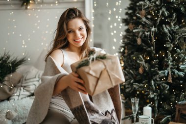 Rahat kış kıyafetleri içinde gülümseyen güzel bir kadın kameraya bir hediye kutusu uzatıyor, bir Noel hediyesi hazırlıyor, kış tatilini kutluyor.
