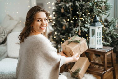 Rahat kış kıyafetleri içinde gülümseyen güzel bir kadın kameraya bir hediye kutusu uzatıyor, bir Noel hediyesi hazırlıyor, kış tatilini kutluyor.