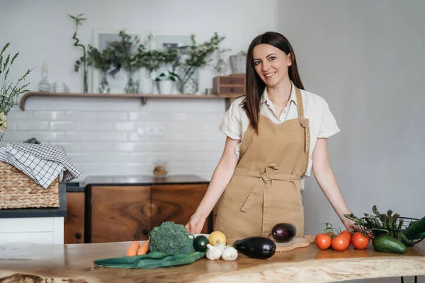 Όμορφη νεαρή μελαχρινή γυναίκα προετοιμάζει στην κουζίνα σύμφωνα με μια συνταγή από ένα σημειωματάριο ένα υγιεινό γεύμα από φρέσκα βιολογικά λαχανικά κοιτάζει την κάμερα — Φωτογραφία Αρχείου