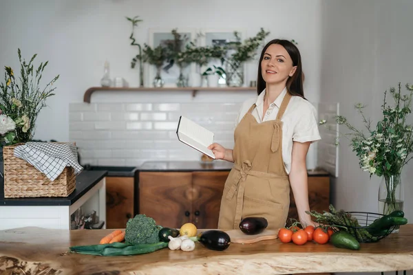 Bela jovem morena mulher se prepara na cozinha de acordo com uma receita de um notebook uma refeição saudável de vegetais orgânicos frescos olha para a câmera — Fotografia de Stock