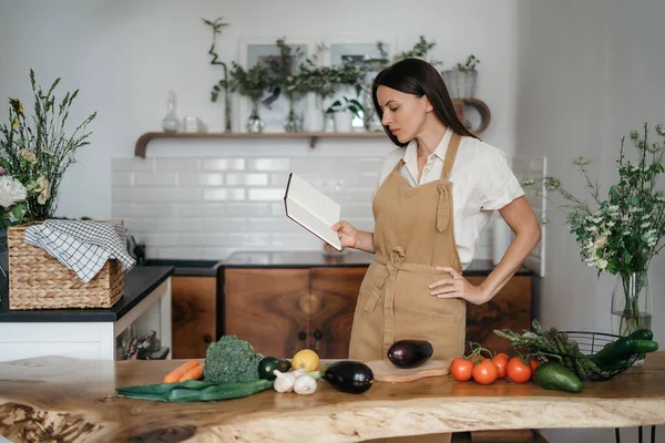 Bela jovem morena prepara uma refeição saudável de vegetais orgânicos frescos na cozinha de acordo com uma receita de um caderno — Fotografia de Stock