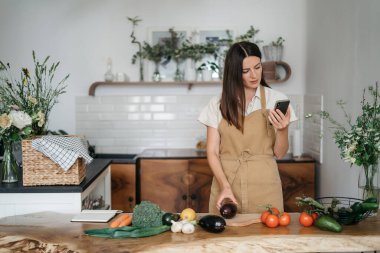 Evdeki mutfakta sağlıklı yiyeceklerle dikilen genç bir kadın telefonu kullanıyor. Cihazları ve sağlıklı gıda konseptiyle modern yaşam tarzı.