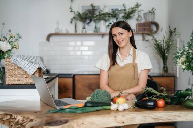 Genç bir kadın mutfakta dizüstü bilgisayar kullanıyor ve sağlıklı vegan sebzeler hazırlıyor. İnternette aşçılık eğitimi
