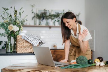 Genç bir kadın mutfakta dizüstü bilgisayar kullanıyor ve sağlıklı vegan sebzeler hazırlıyor. İnternette aşçılık eğitimi