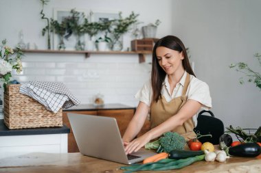 Genç bir kadın mutfakta dizüstü bilgisayar kullanıyor ve sağlıklı vegan sebzeler hazırlıyor. İnternette aşçılık eğitimi