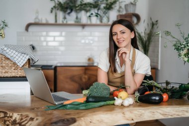 Genç bir kadın mutfakta dizüstü bilgisayar kullanıyor ve sağlıklı vegan sebzeler hazırlıyor. İnternette aşçılık eğitimi