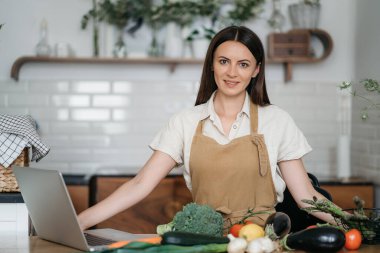 Genç bir kadın mutfakta dizüstü bilgisayar kullanıyor ve sağlıklı vegan sebzeler hazırlıyor. İnternette aşçılık eğitimi