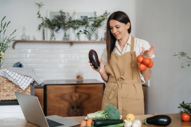 Genç bir kadın mutfakta dizüstü bilgisayar kullanıyor ve sağlıklı vegan sebzeler hazırlıyor. İnternette aşçılık eğitimi