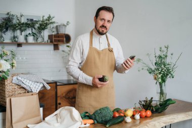 Mutlu genç adam romantik bir akşam yemeği hazırlıyor sebze tarifi arıyor diyet yemek kitabı uygulaması akıllı telefon kullanıyor, elinde telefonla telefon tutuyor mutfak içinde sağlıklı vejetaryen yemeği salatası pişiriyor.