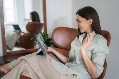 Gülümseyen genç esmer bir sandalyede rahatça oturur ve bir tablet kullanır. Günlük giysiler içinde mutlu bir bayan boş zamanlarını kablosuz bilgisayarla evde geçiriyor..