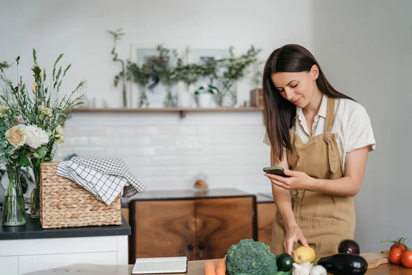 Jovem usando telefone enquanto está de pé com comida saudável na cozinha em casa. Estilo de vida moderno com gadgets e conceito de comida saudável — Fotografia de Stock