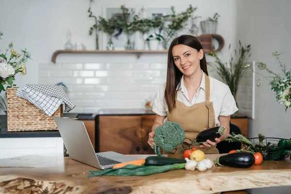 Uma jovem mulher está usando um laptop na cozinha preparando legumes veganos saudáveis. Treinamento de culinária online — Fotografia de Stock