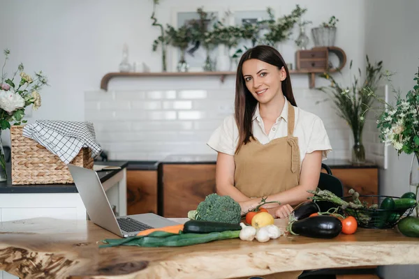 Uma jovem mulher está usando um laptop na cozinha preparando legumes veganos saudáveis. Treinamento de culinária online — Fotografia de Stock