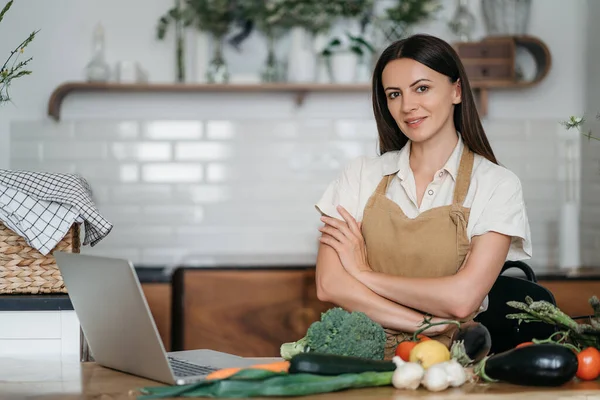 Uma jovem mulher está usando um laptop na cozinha preparando legumes veganos saudáveis. Treinamento de culinária online — Fotografia de Stock