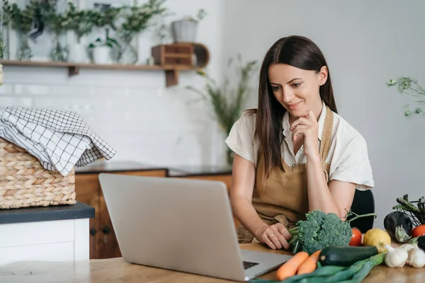 Uma jovem mulher está usando um laptop na cozinha preparando legumes veganos saudáveis. Treinamento de culinária online — Fotografia de Stock