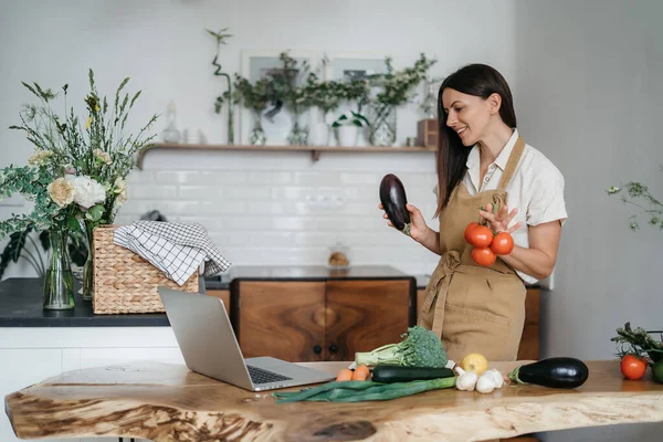 Genç bir kadın mutfakta dizüstü bilgisayar kullanıyor ve sağlıklı vegan sebzeler hazırlıyor. İnternette aşçılık eğitimi