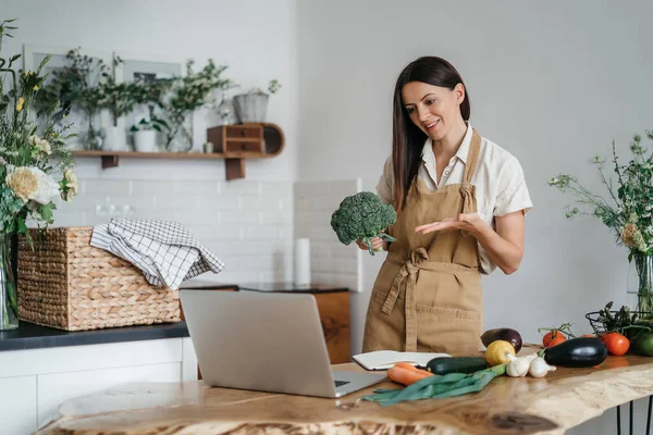 Uma jovem mulher está usando um laptop na cozinha preparando legumes veganos saudáveis. Treinamento de culinária online — Fotografia de Stock