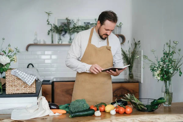 Mutlu genç adam romantik bir akşam yemeği hazırlıyor sebze tarifi arıyor diyet yemek kitabı uygulaması akıllı telefon kullanıyor, elinde telefonla telefon tutuyor mutfak içinde sağlıklı vejetaryen yemeği salatası pişiriyor.