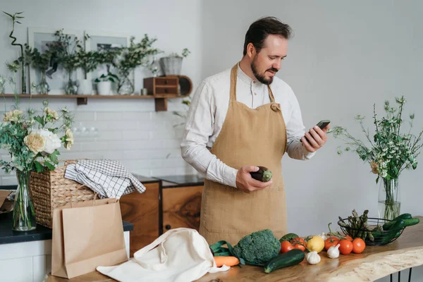 Mutlu genç adam romantik bir akşam yemeği hazırlıyor sebze tarifi arıyor diyet yemek kitabı uygulaması akıllı telefon kullanıyor, elinde telefonla telefon tutuyor mutfak içinde sağlıklı vejetaryen yemeği salatası pişiriyor.