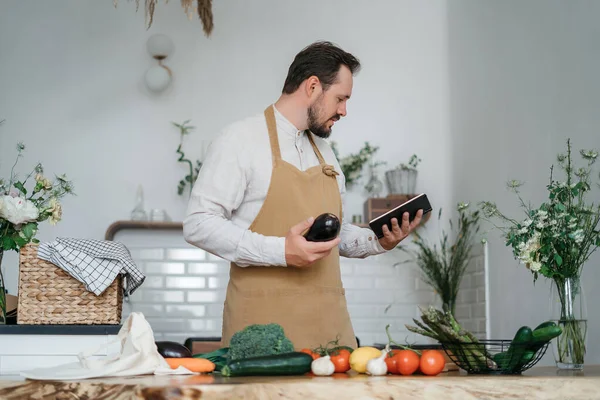 Mutlu genç adam romantik bir akşam yemeği hazırlıyor sebze tarifi arıyor diyet yemek kitabı uygulaması akıllı telefon kullanıyor, elinde telefonla telefon tutuyor mutfak içinde sağlıklı vejetaryen yemeği salatası pişiriyor.