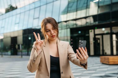 beautiful blonde woman takes a selfie in the city dressed in a jacket on the background of buildings. video call by phone