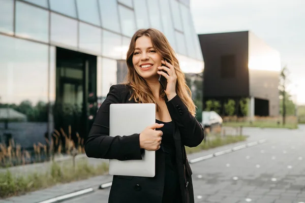 Portrait of a beautiful blonde business woman in a jacket and glasses smiling while talking on the phone on the street