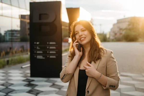 Portrait of a beautiful blonde business woman in a jacket smiling talking on the phone in the street at sunset