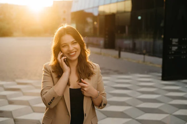 Portrait of a beautiful blonde business woman in a jacket smiling talking on the phone in the street at sunset