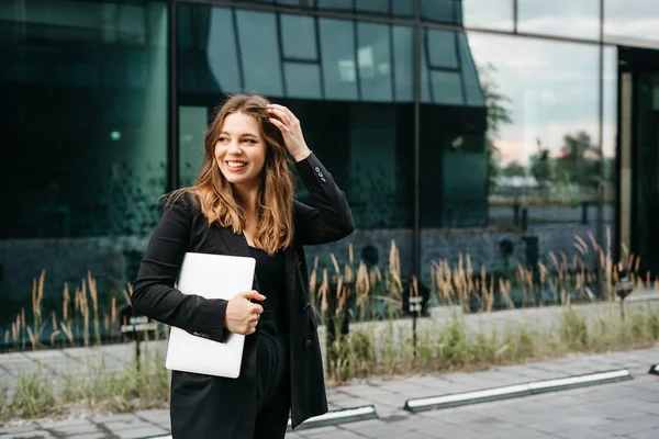 smiling positive business woman standing with laptop near business center, confident small business owner looking at camera, office worker in black jacket formal suit