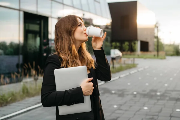 smiling positive business woman standing with laptop near business center, drinking coffee during lunch break, office worker in black jacket formal suit