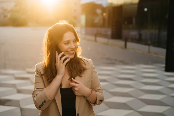 Portrait of a beautiful blonde business woman in a jacket smiling talking on the phone in the street at sunset