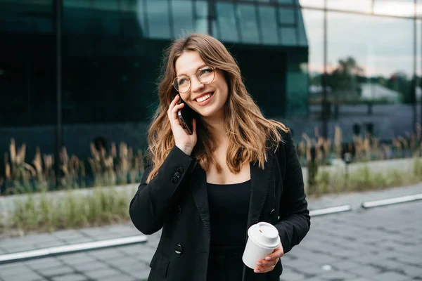 Portrait of a beautiful blonde business woman in a jacket and glasses smiling while talking on the phone on the street
