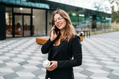 Lifestyle portrait of a young and cheerful woman standing with phone and coffee cup outdoors. Urban business travel and transportation concept