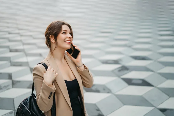 Lifestyle portrait of young and cheerful woman standing with phone and backpack outdoors. City business travel and transport concept