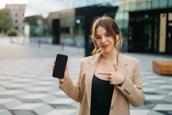 Young stylish woman shows mobile phone while standing relaxed in modern buildings business center. Phone with black screen to copy and paste