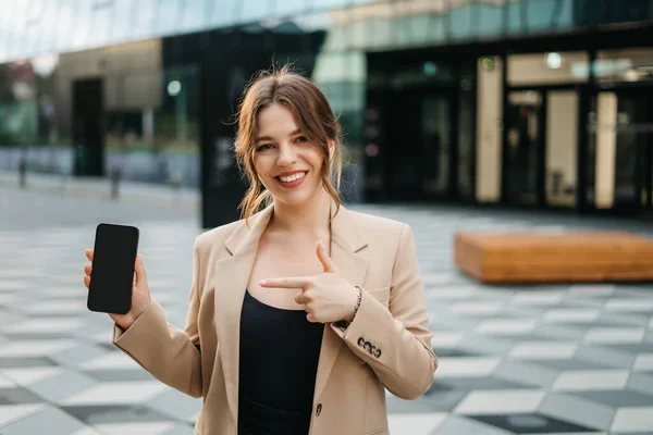 Young stylish woman shows mobile phone while standing relaxed in modern buildings business center. Phone with black screen to copy and paste