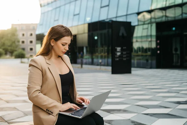 Happy young woman blonde office worker typing on laptop while sitting outdoors in the city, business woman or female freelancer answering emails, working outdoors, cityscape in the background.