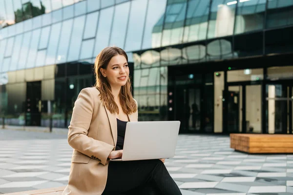 Happy young woman blonde office worker typing on laptop while sitting outdoors in the city, business woman or female freelancer answering emails, working outdoors, cityscape in the background.